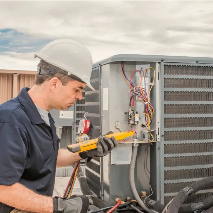 HVAC technician in a hard hat testing an outdoor air conditioner with a multimeter